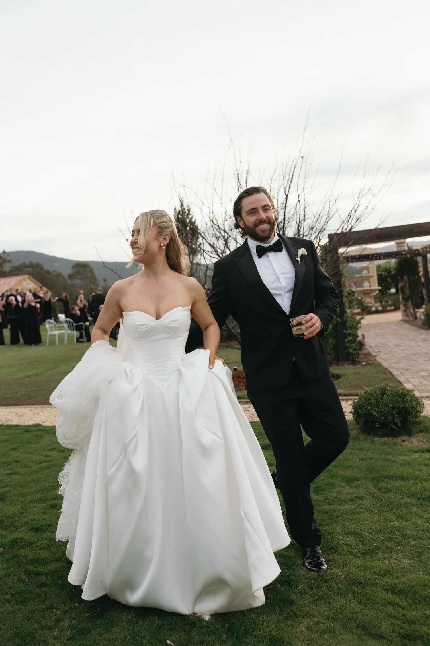 Bride in white strapless gown and groom in black tuxedo pose together outside an elegant venue with lush greenery.