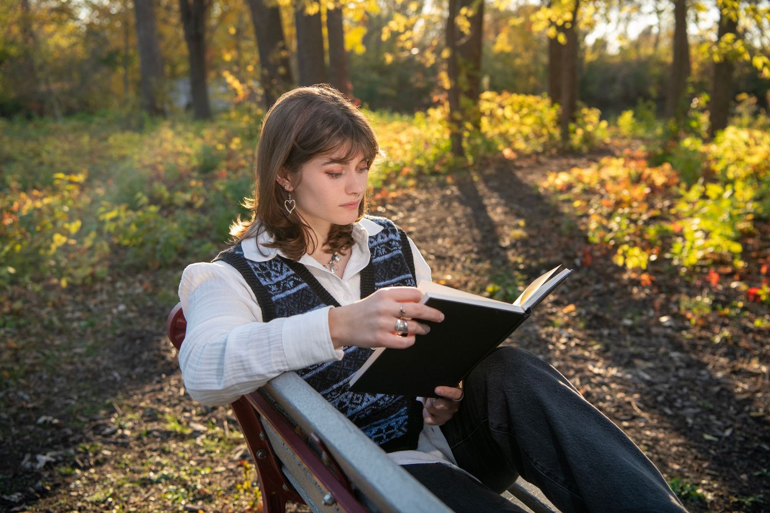 Young woman reading a book on a park bench surrounded by autumn foliage in golden sunlight.