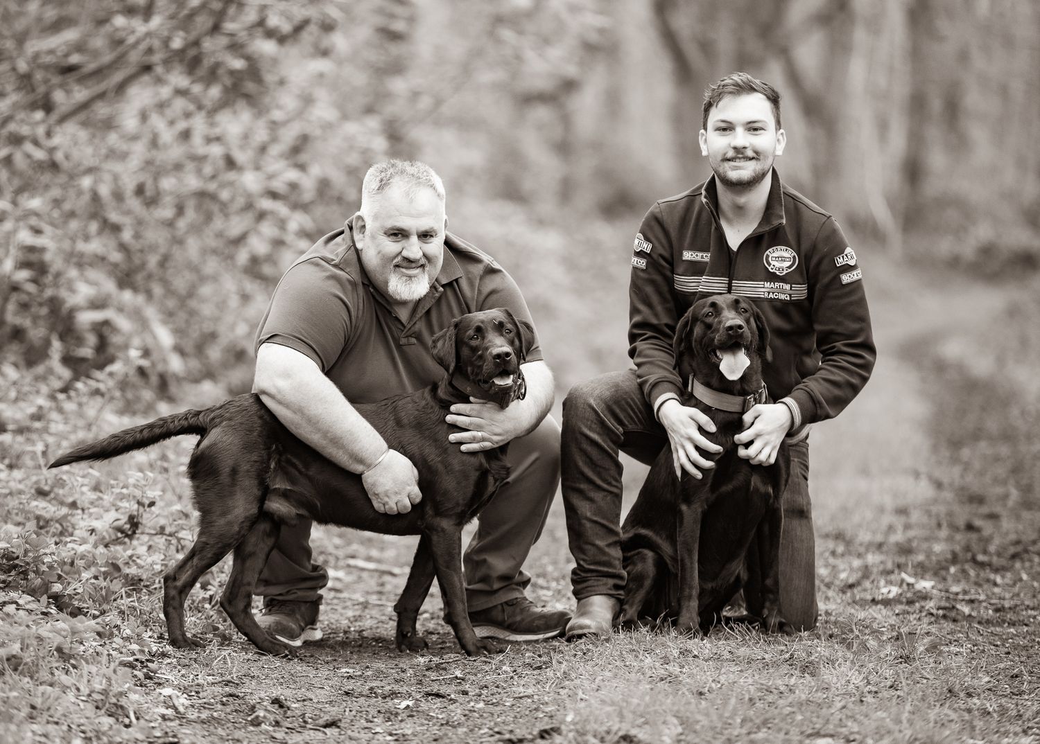 Two men kneeling outdoors with two black Labrador dogs in a black and white portrait photo.