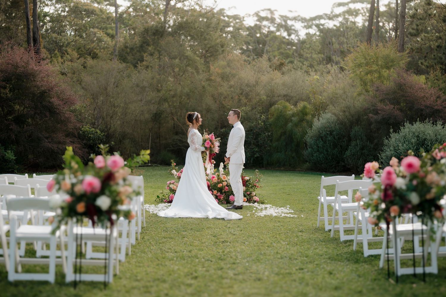 Bride and groom stand facing each other at an outdoor garden wedding ceremony with white chairs and pink floral arrangements.
