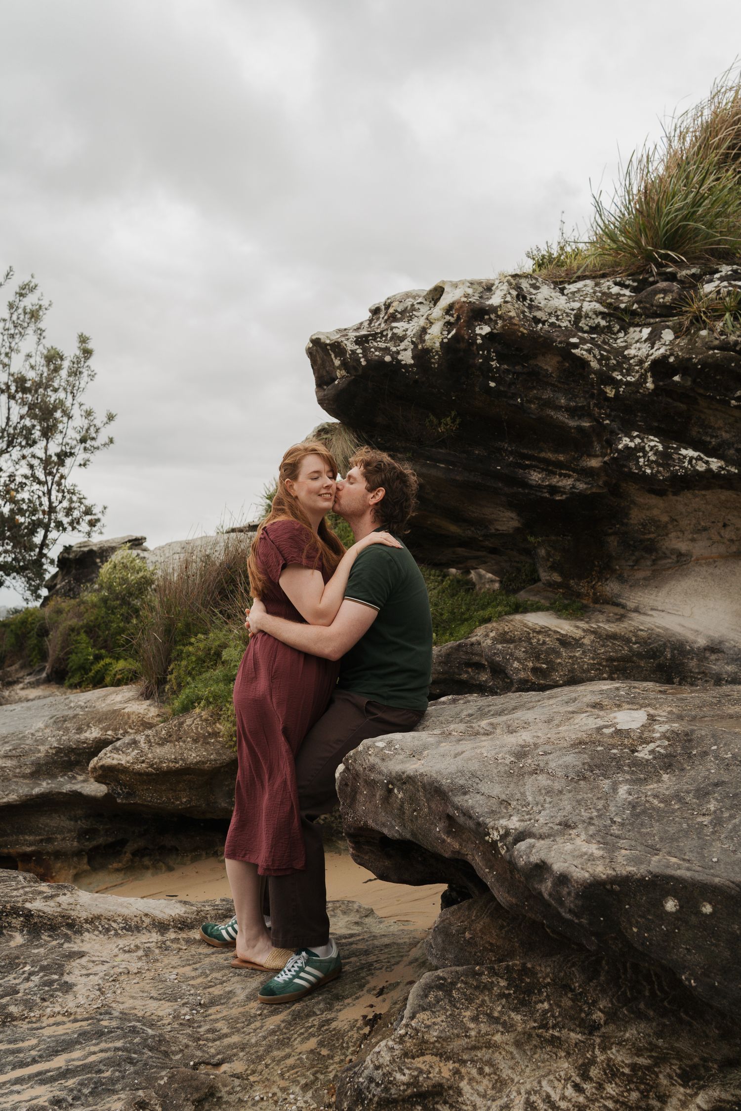 A couple shares a romantic kiss while embracing on rocky terrain under a cloudy sky outdoors engagement photoshoot in sydney milk beach