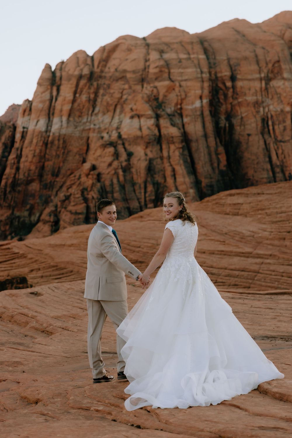 Bride in flowing white gown holds grooms hand on red sandstone cliffs at outdoor wedding.