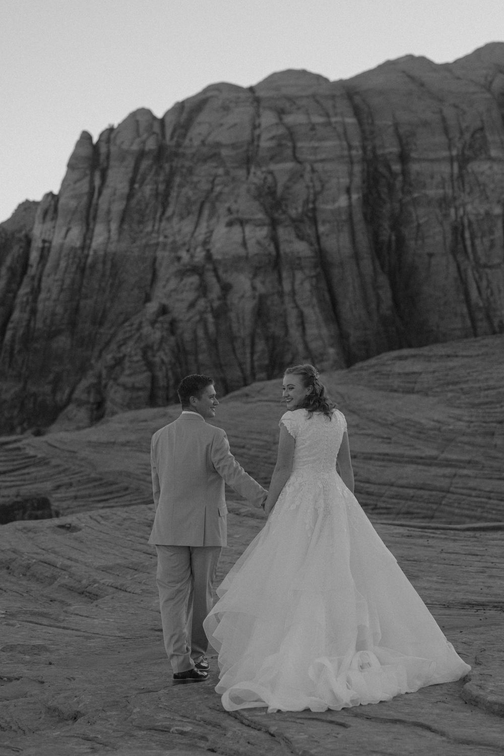Black and white photo of bride and groom holding hands walking on red rock desert landscape.