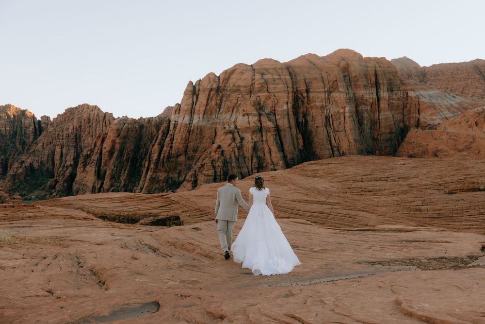 Bride and groom walking hand in hand toward dramatic red sandstone canyon walls at sunset.