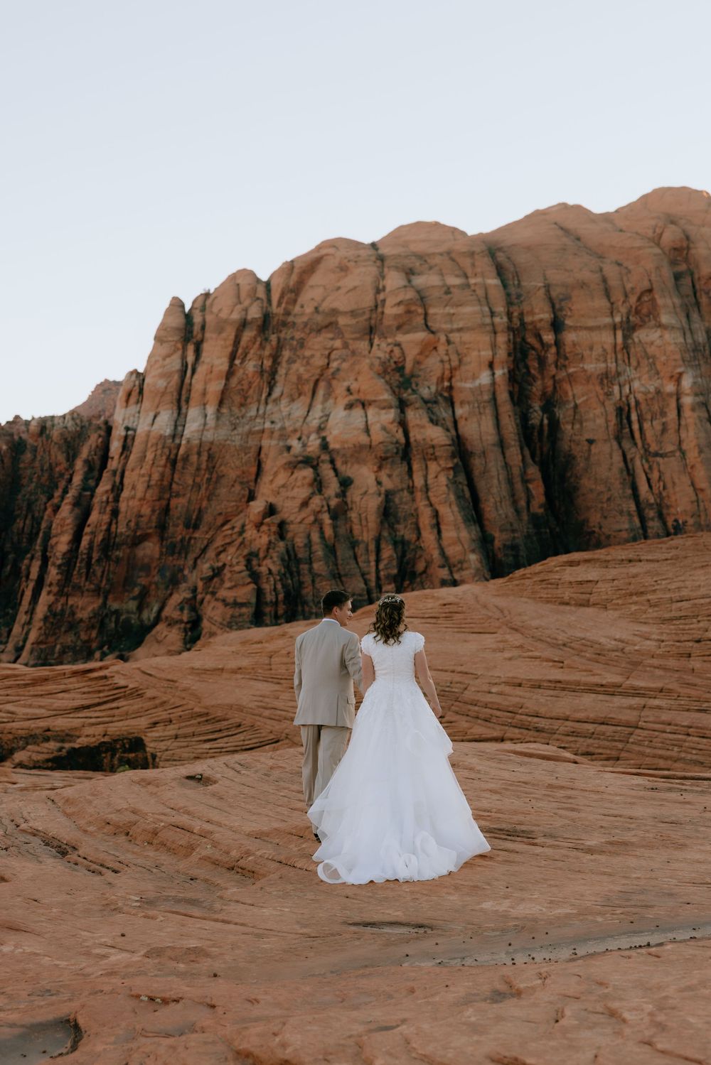 Bride and groom walk hand in hand toward stunning red rock canyon walls at golden hour.
