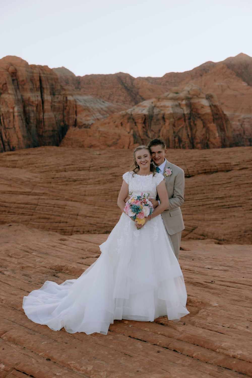 Bride and groom pose together on red rock desert landscape, bride holding colorful bouquet in flowing white gown.