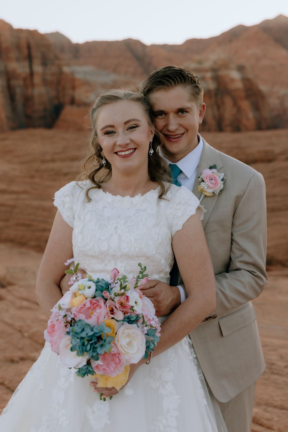 Smiling bride and groom posing with colorful bouquet against red rock canyon wedding backdrop.