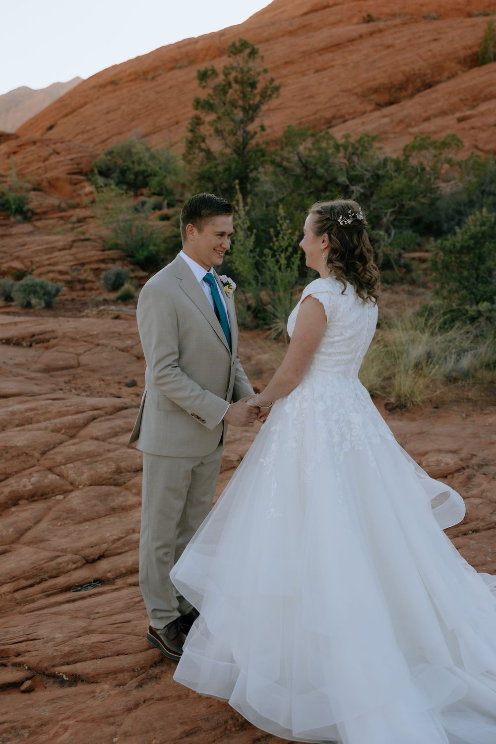 Bride in layered white gown and groom in tan suit hold hands and share a glance on red rock desert terrain.