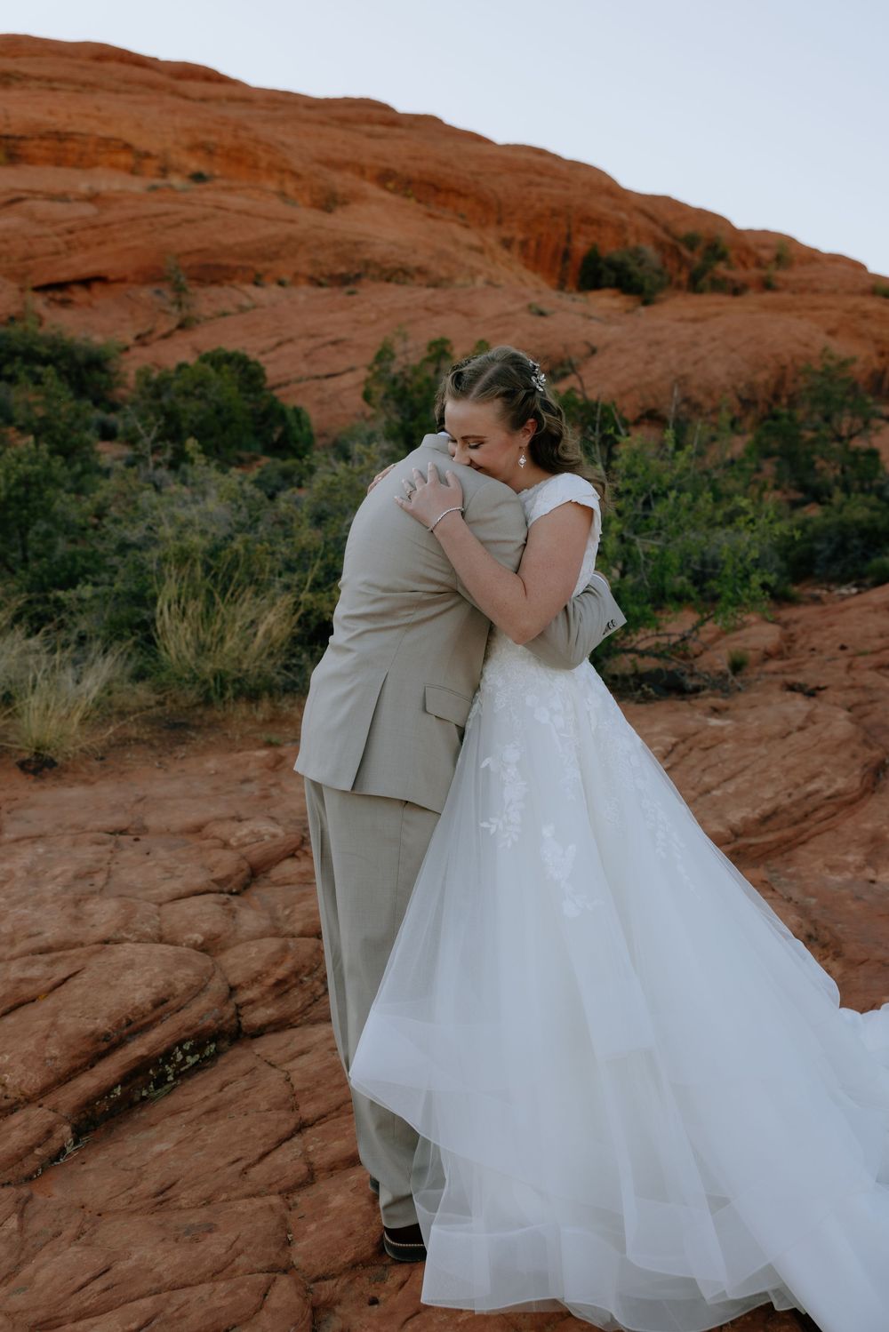 Bride and groom share heartfelt embrace among red sandstone rocks and desert greenery on wedding day.