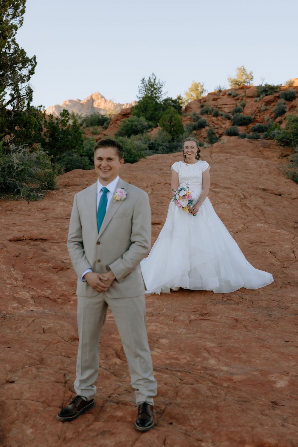 Groom in tan suit with teal tie stands before bride in white gown on red rock desert landscape.