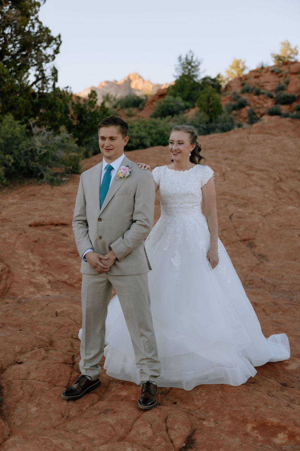 Bride in white lace gown and groom in tan suit pose together smiling on red sandstone with desert cliffs behind them.