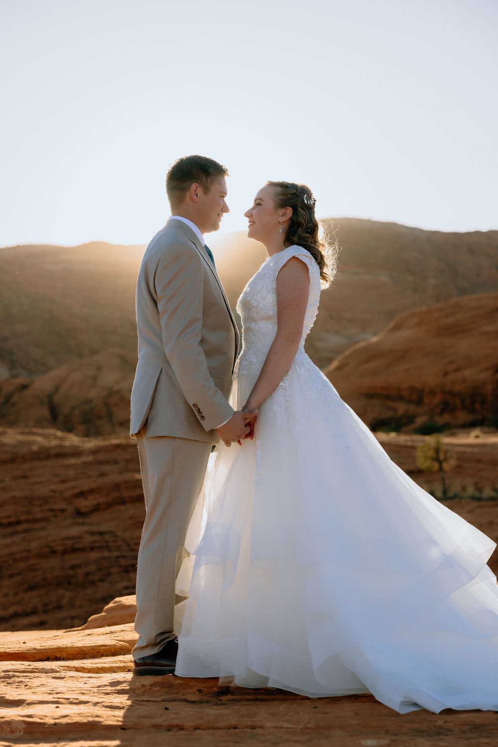 Bride and groom hold hands facing each other at sunset in a red rock desert landscape.