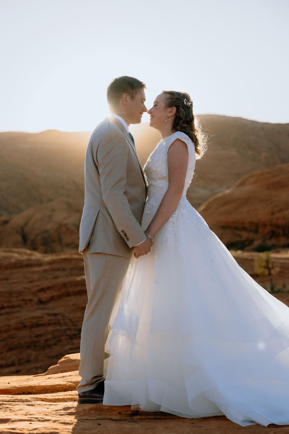Bride and groom hold hands and lean in for a kiss backlit by golden sunset over red rock desert landscape.