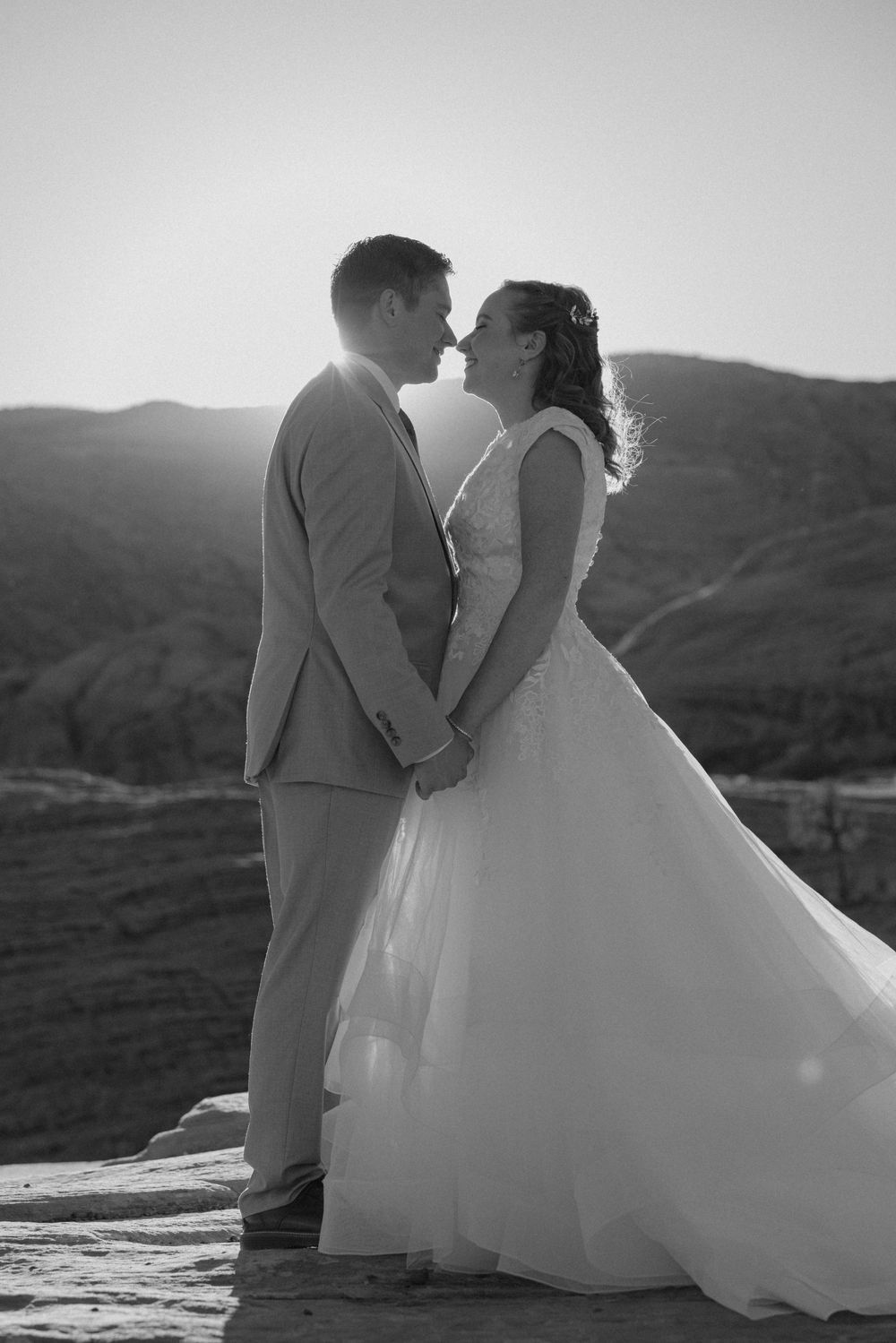 Black and white photo of bride and groom holding hands and sharing a romantic moment at sunset in a desert landscape.