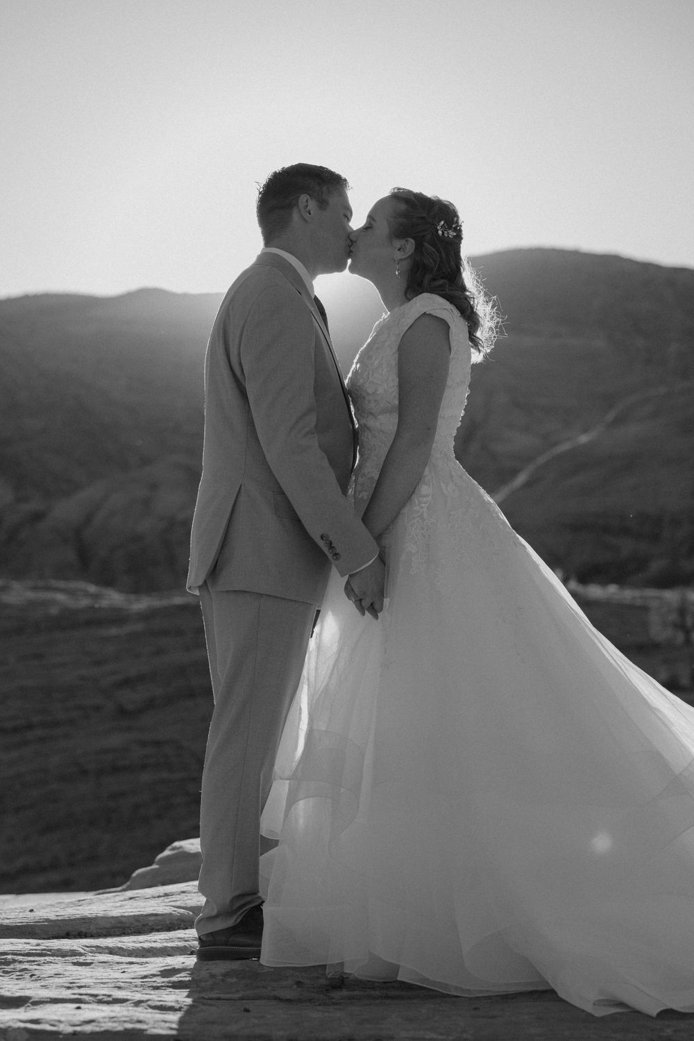 Black and white photo of bride and groom kissing while holding hands with mountain backdrop.