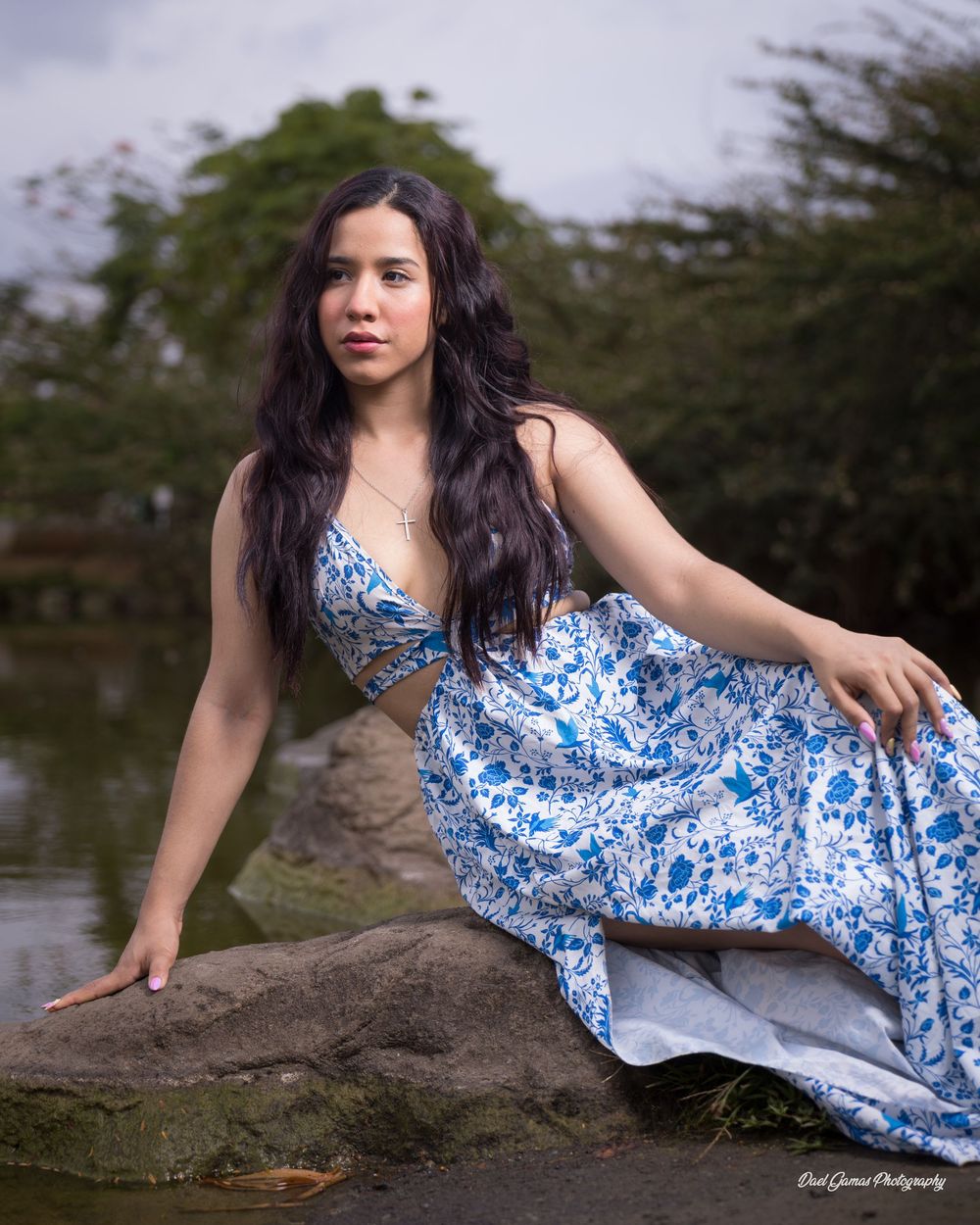 Young woman in blue floral dress poses on rocks by a serene pond surrounded by lush green trees.