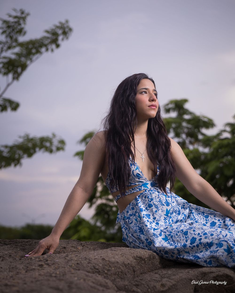Young woman in blue floral dress sitting on rocks outdoors at dusk with trees in background.