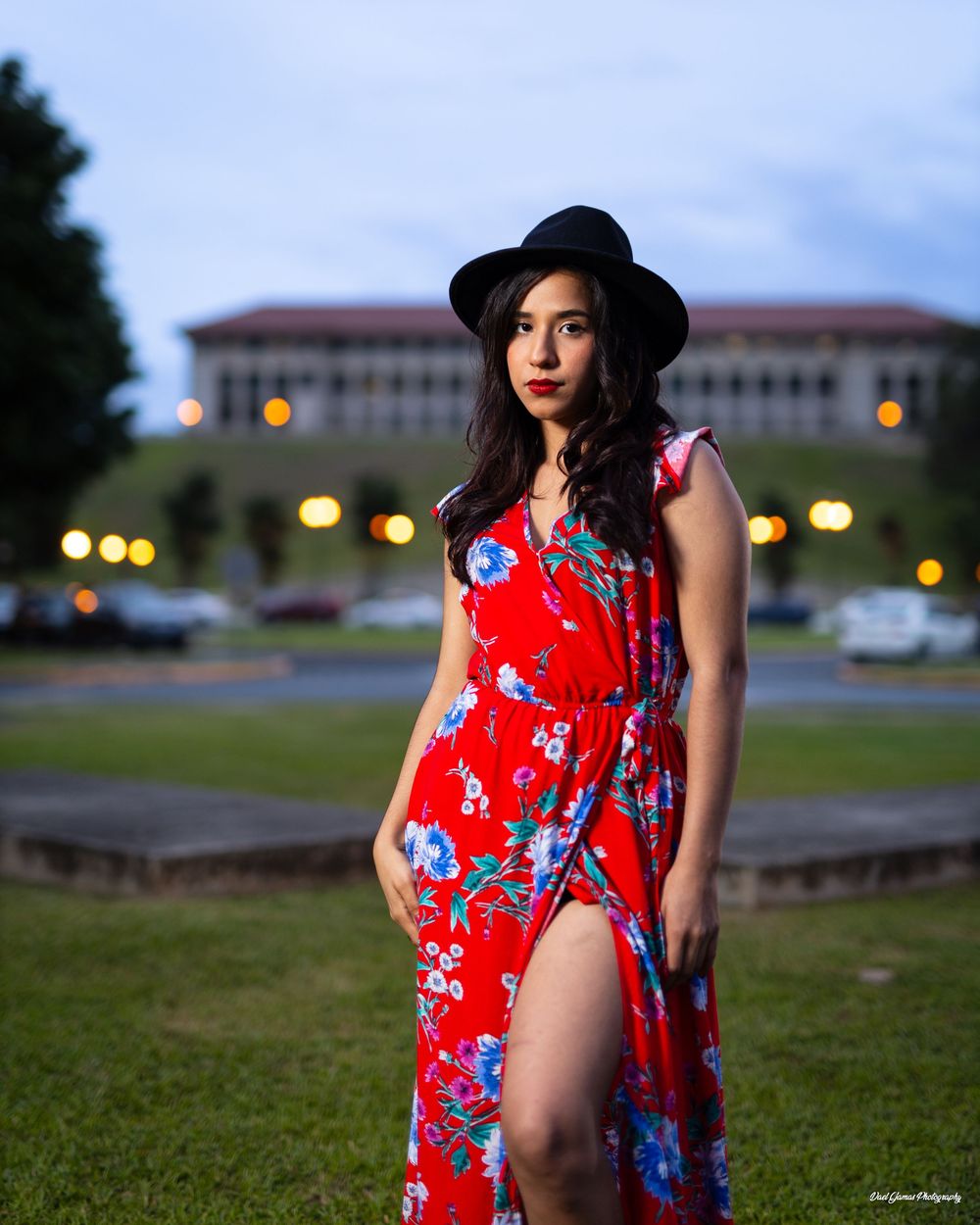 Woman in red floral dress and black hat posing outdoors at dusk near a historic building.