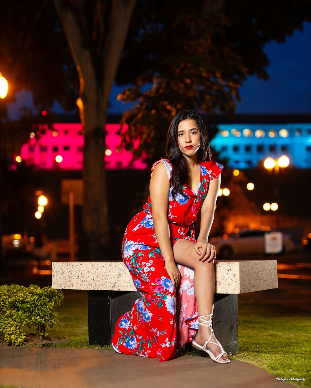 Woman in red floral dress seated on bench at night with colorful city lights in background.