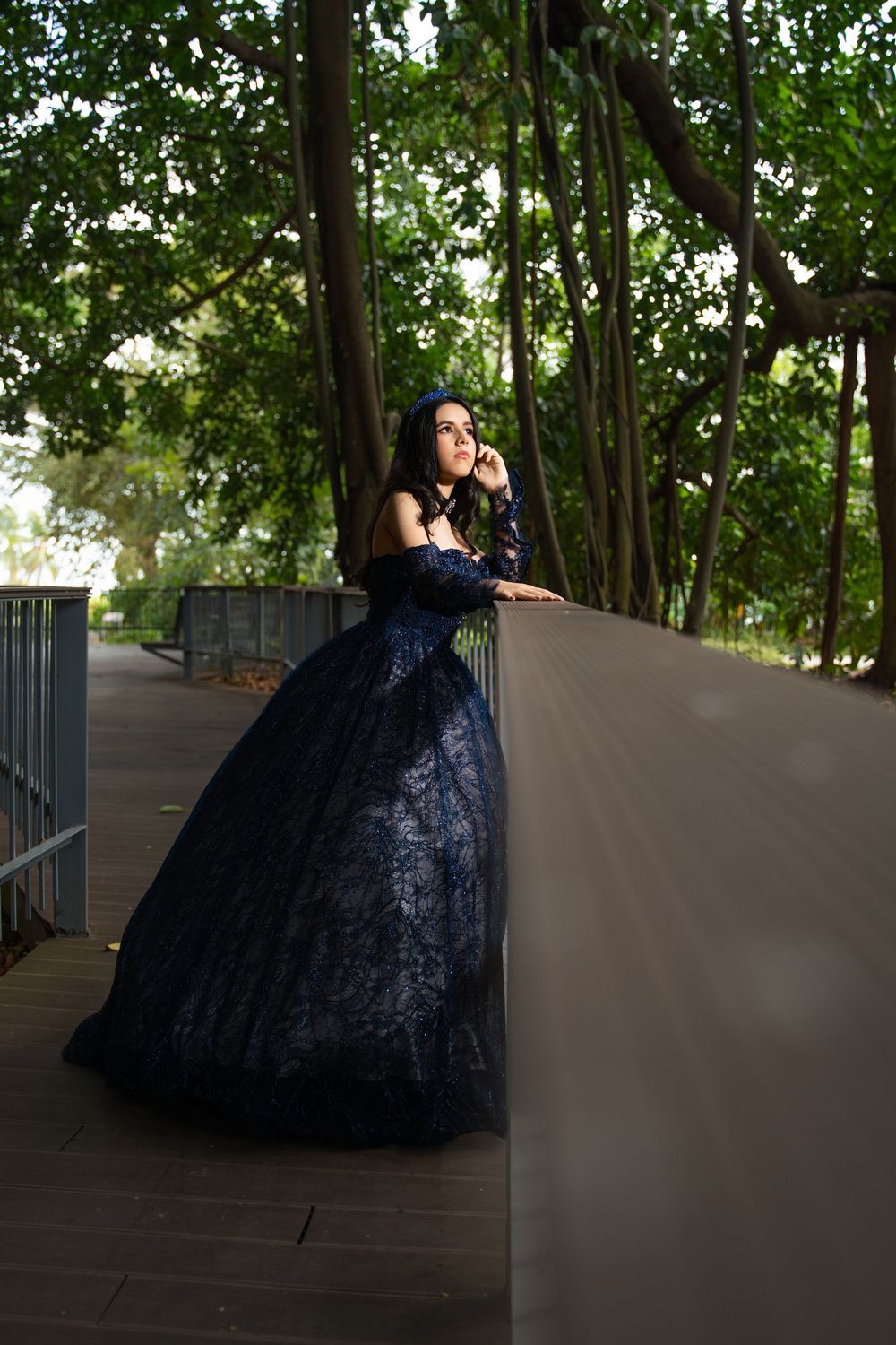 Woman in elegant black lace ballgown posing gracefully on a wooden walkway surrounded by lush green trees.