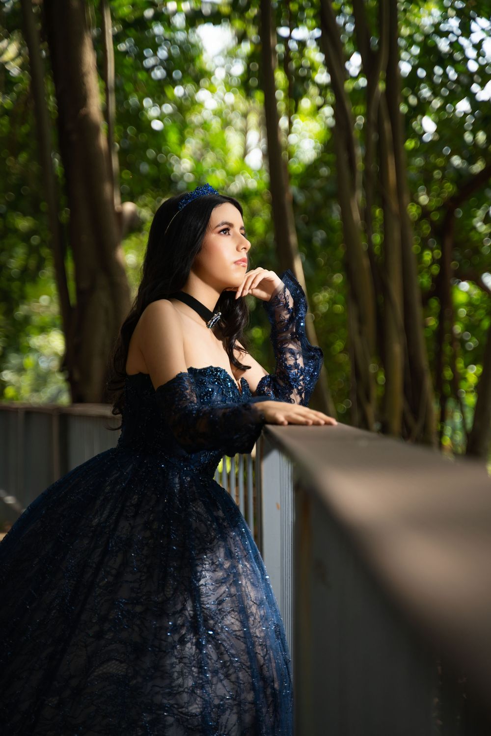 Young woman in elegant navy blue lace ball gown poses on a forest bridge, gazing thoughtfully into the distance.