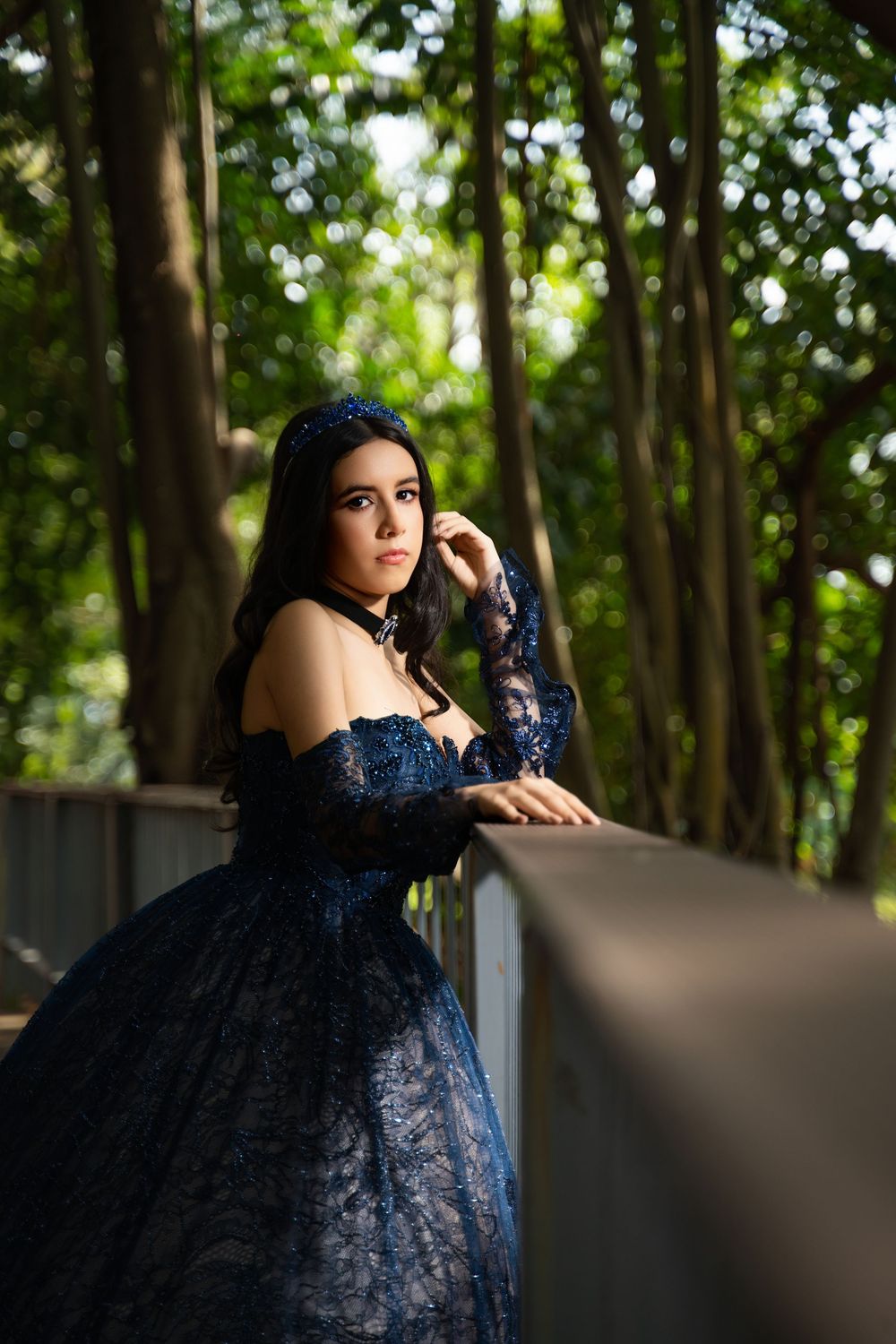 Young woman in elegant dark blue lace ball gown and crown poses gracefully on a bridge surrounded by lush green trees.