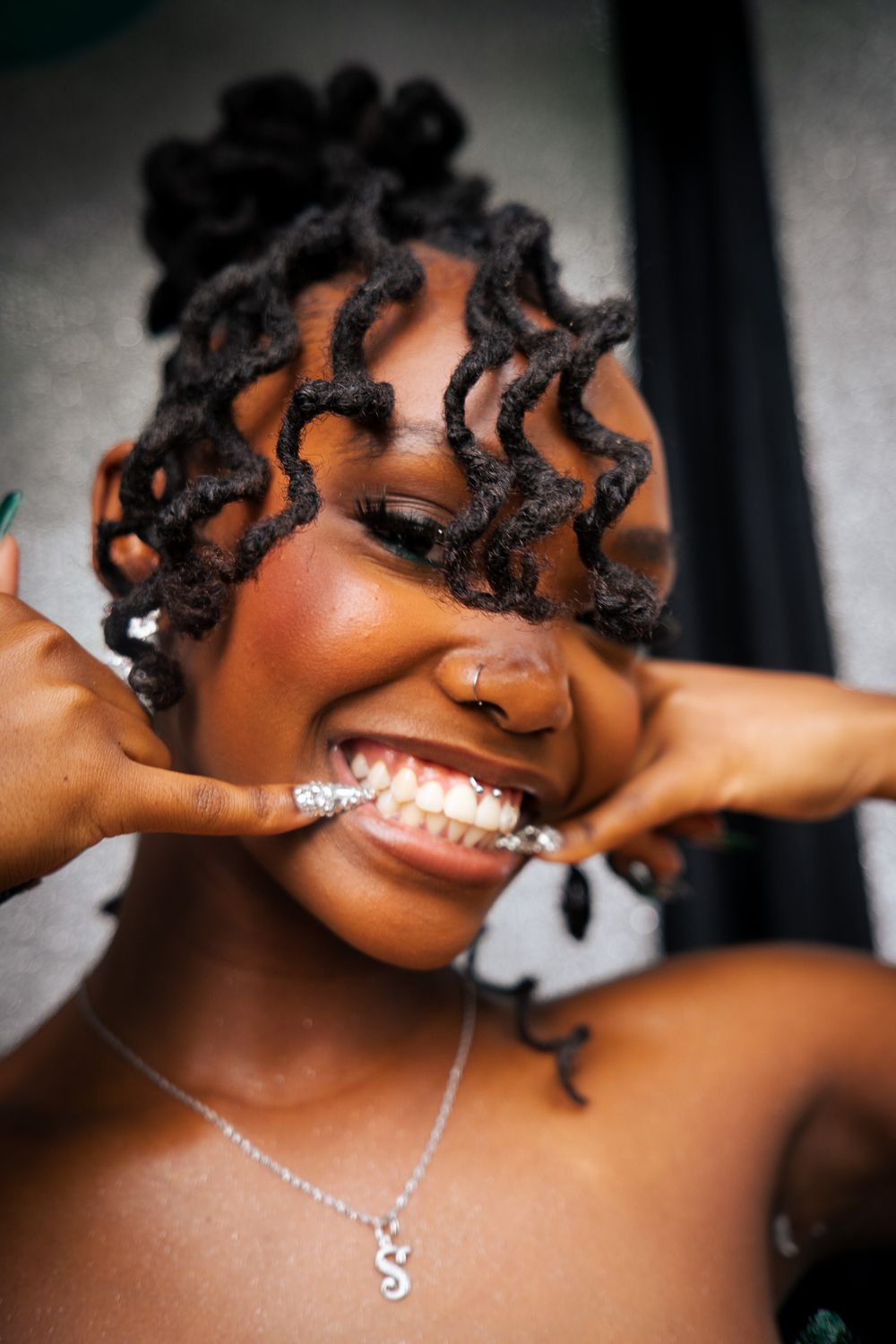 Young woman with twisted locs hairstyle smiling broadly, wearing an S initial necklace and green nails.