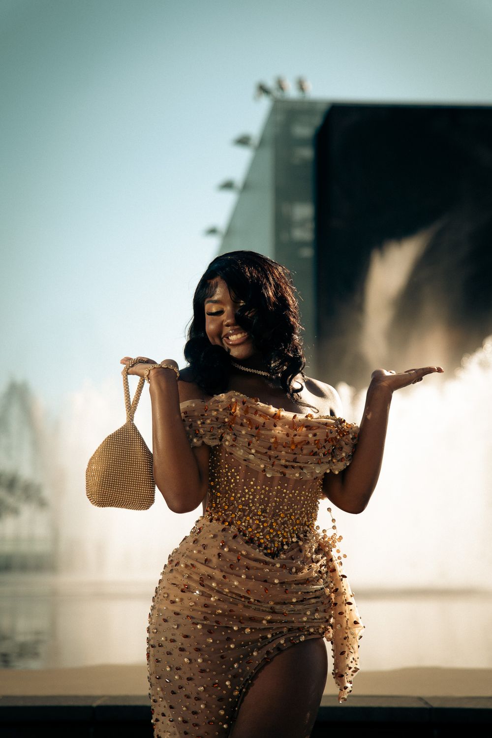 A smiling woman in a gold embellished off-shoulder dress holds a woven bag near a fountain.
