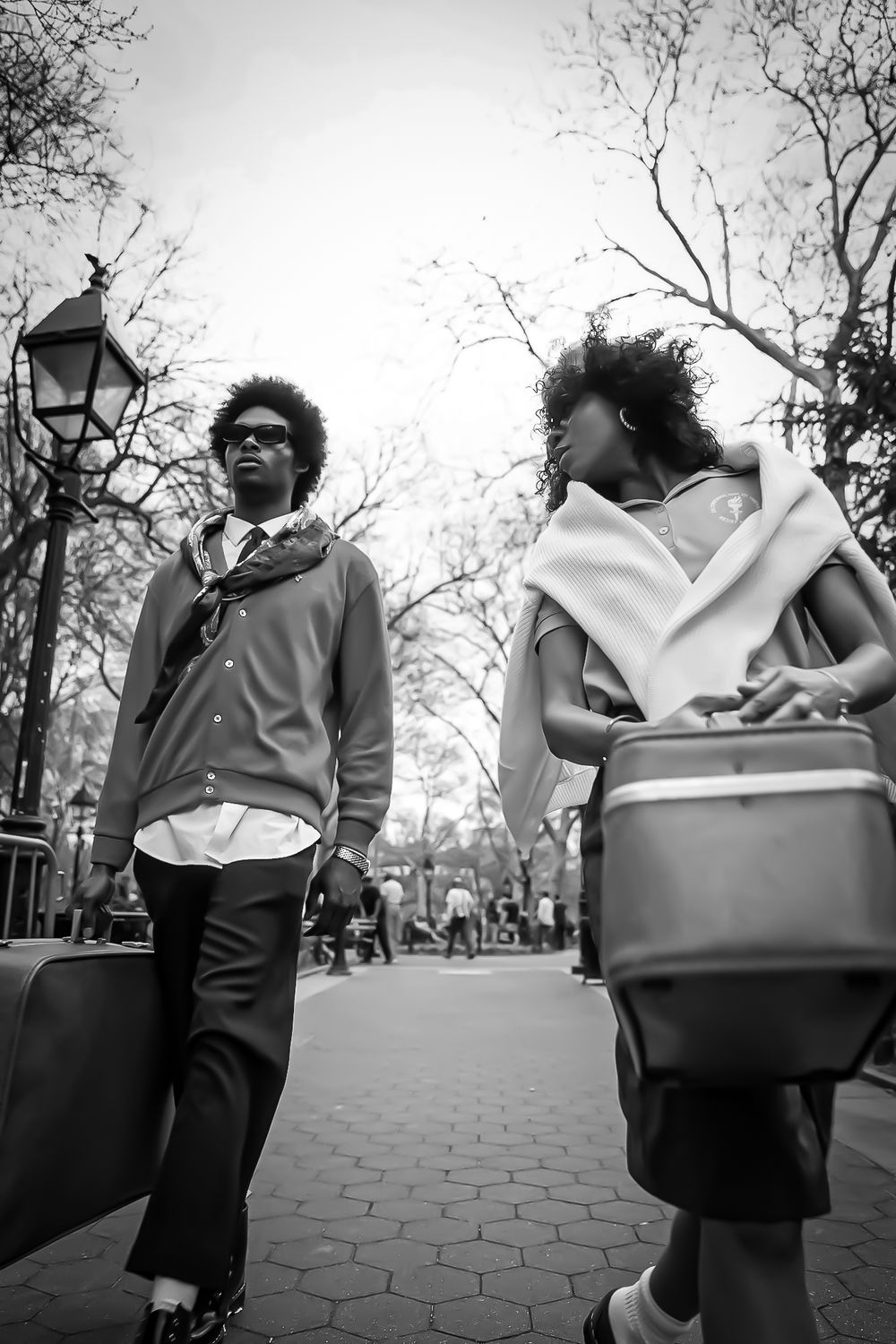 Black and white photo of two young adults walking in a park, carrying bags and a cooler on a tree-lined path.