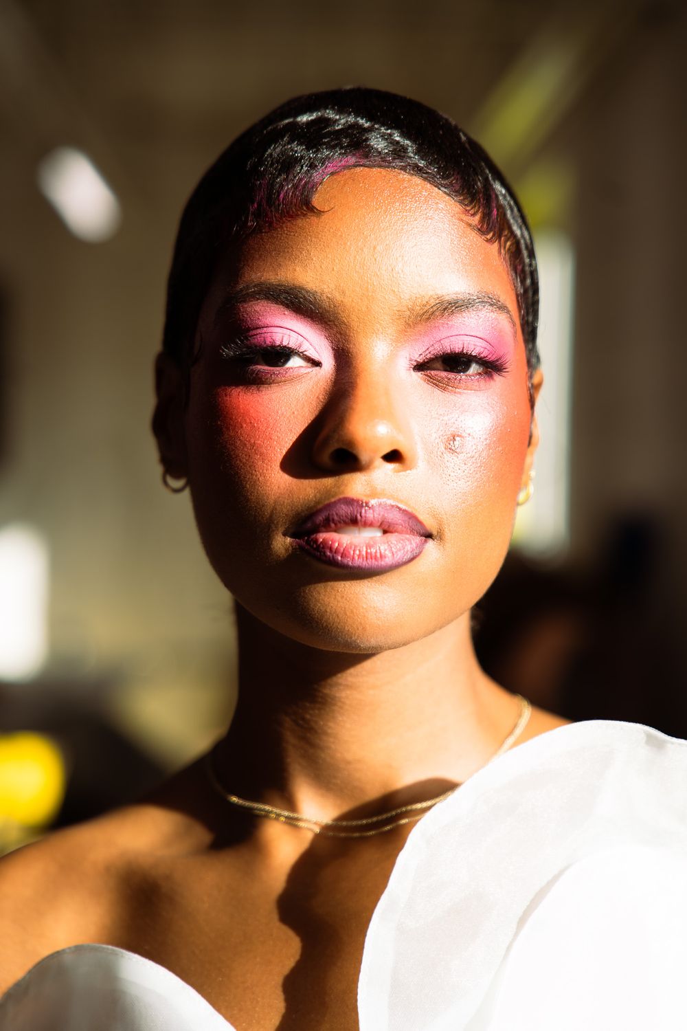 Close-up portrait of a woman with bold pink eye makeup and rosy blush, wearing a white top and gold necklace.