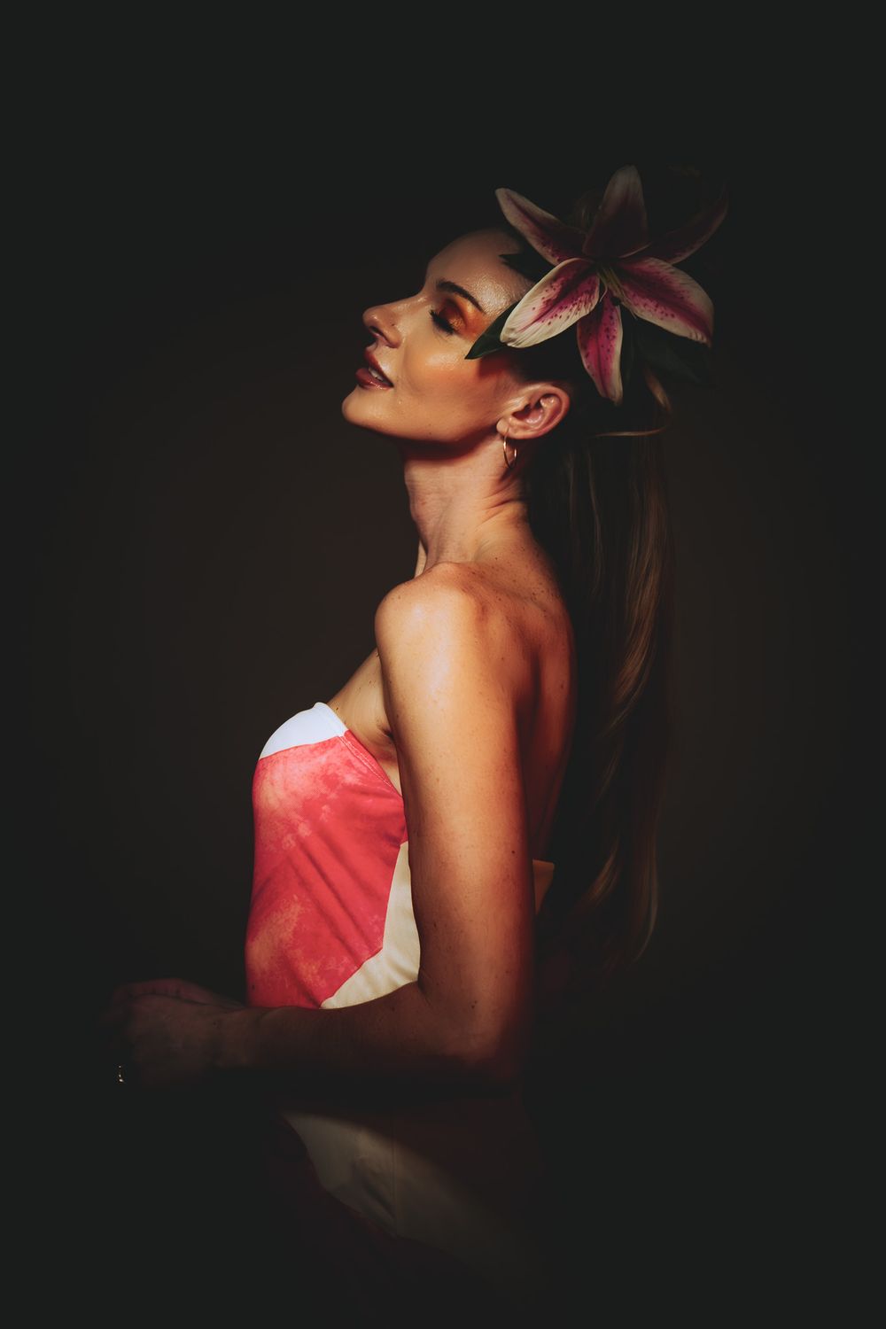 Side profile of a woman with a pink lily flower in her hair wearing a strapless pink top.