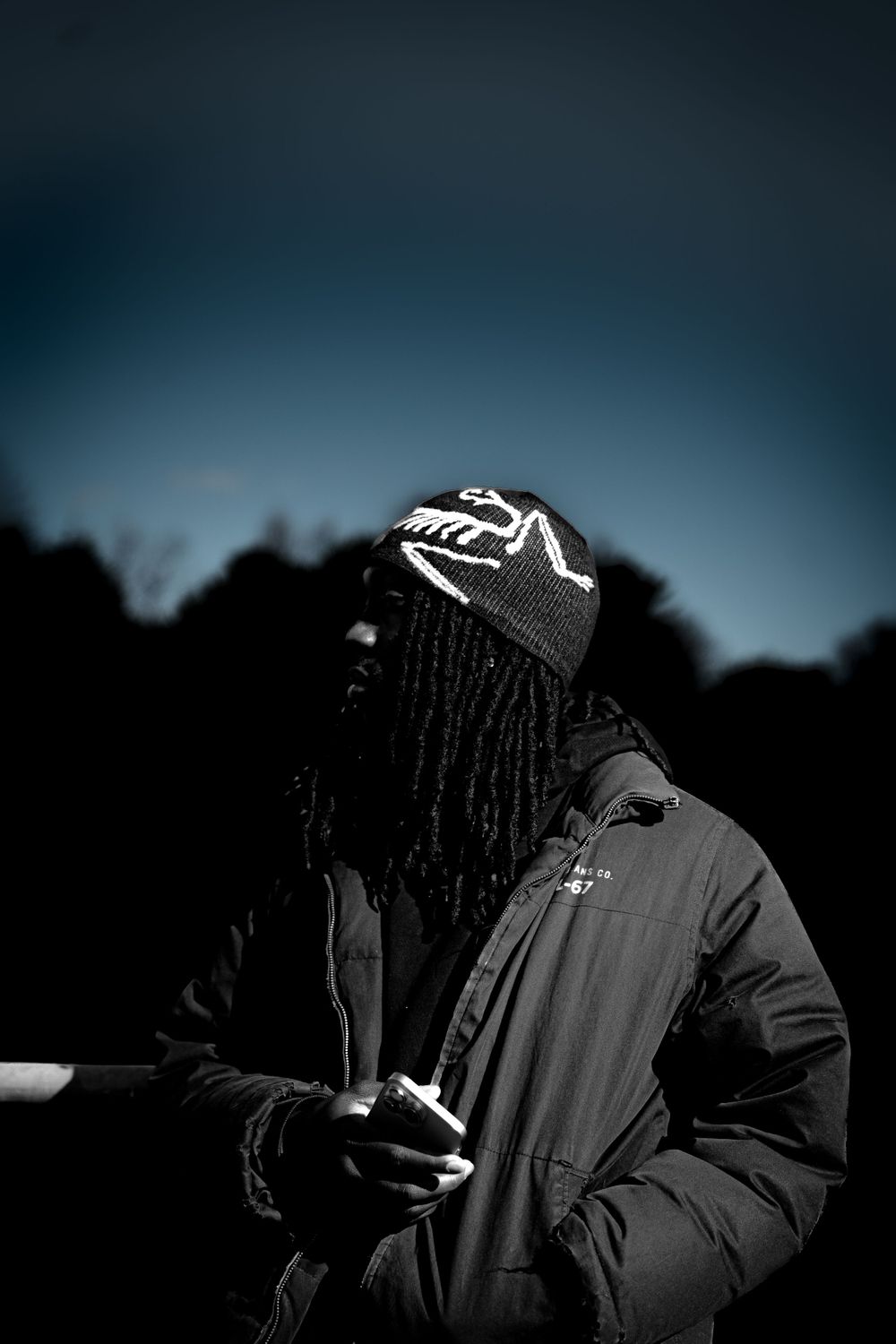 Man with dreadlocks wearing black beanie and jacket holds phone outdoors against dramatic dark blue sky.