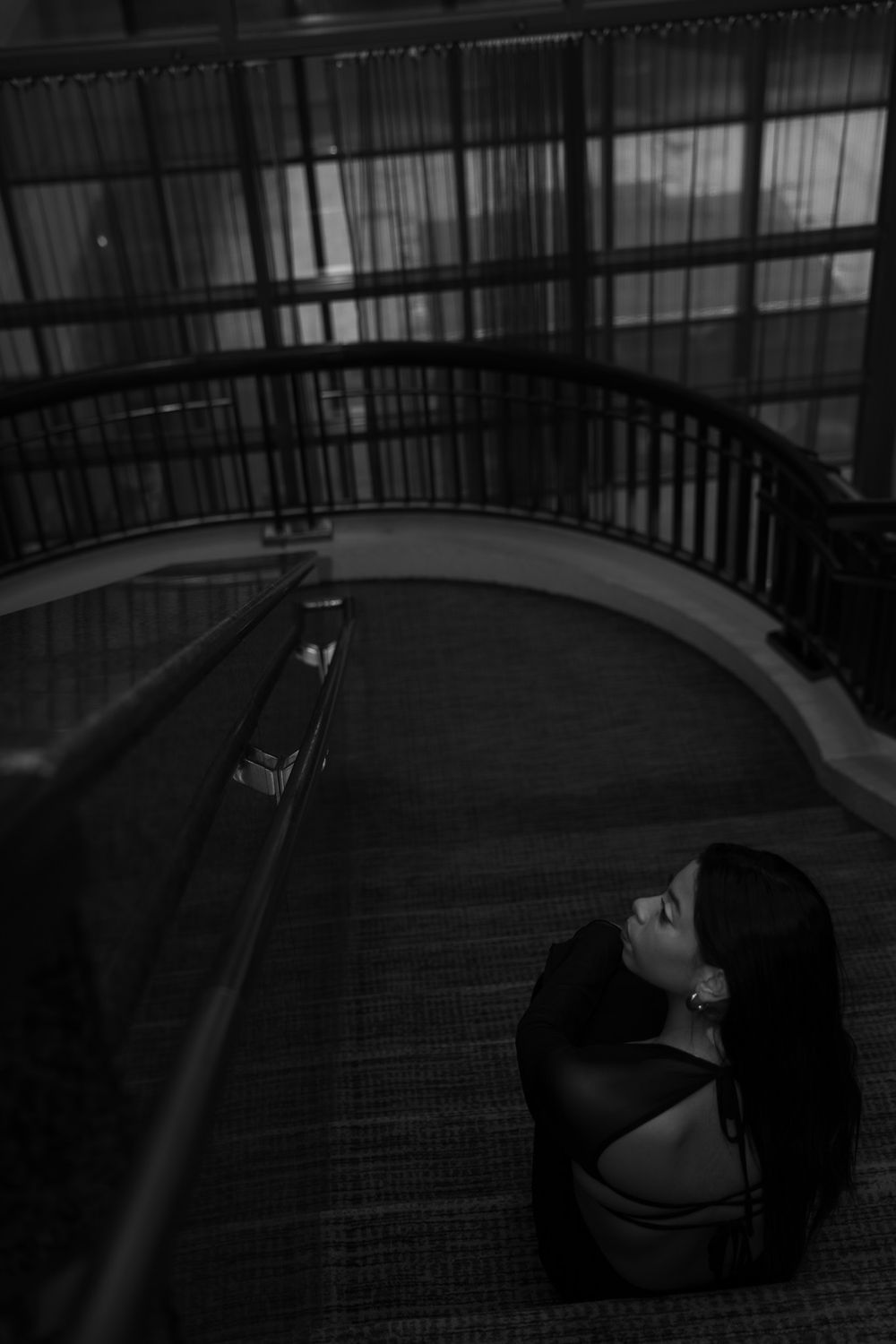 Woman with long dark hair sits on carpeted stairs by curved railing in black and white photo.