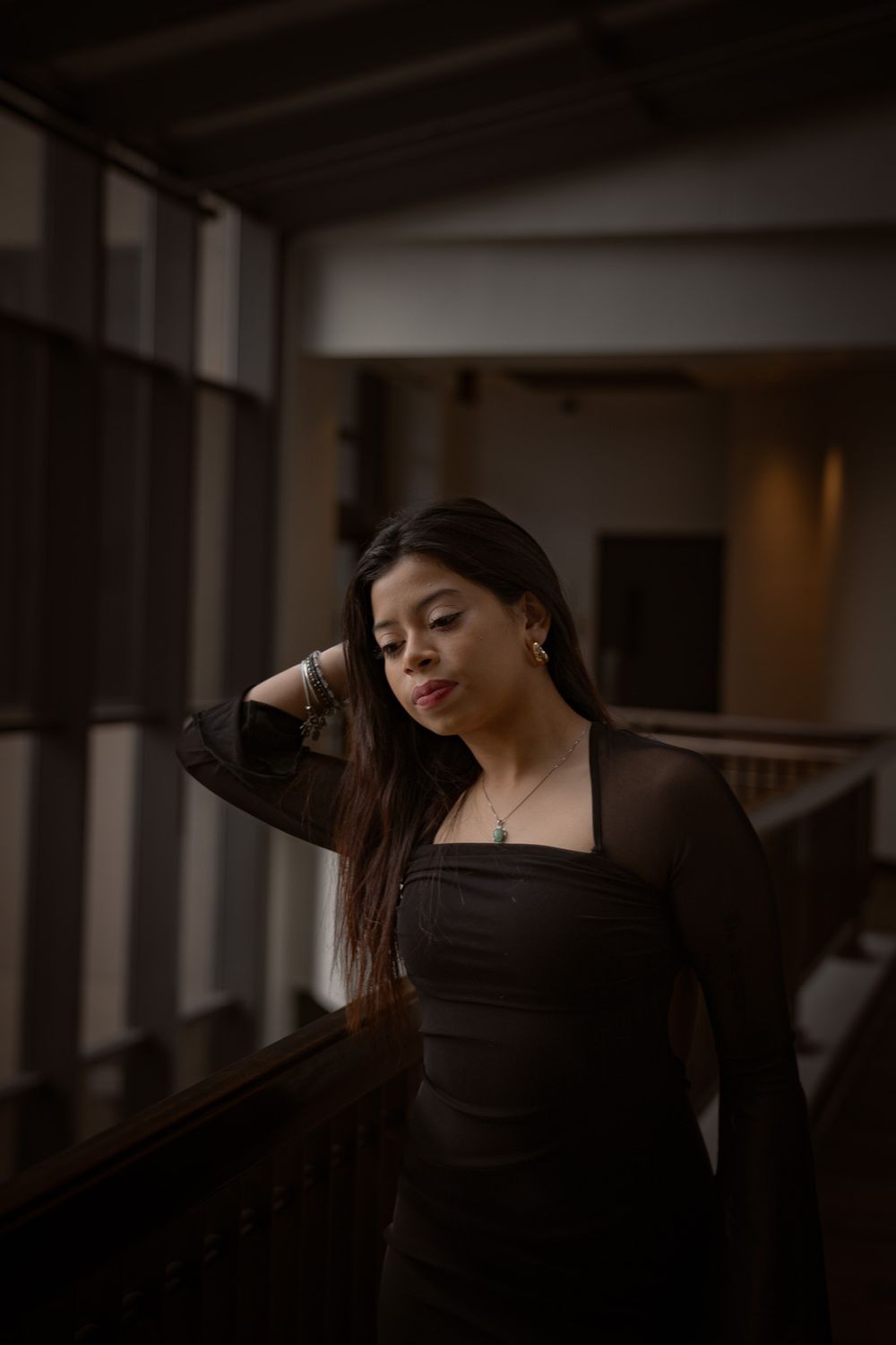Young Asian woman in black mesh dress stands by railing near window in moody indoor setting.