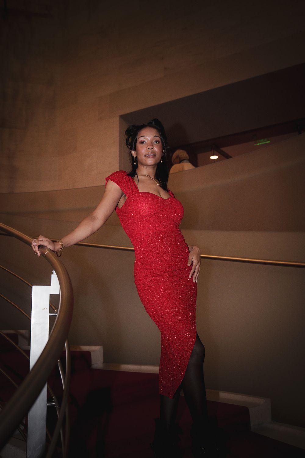 Woman in sparkling red fitted dress posing gracefully on a curved staircase with gold railing.