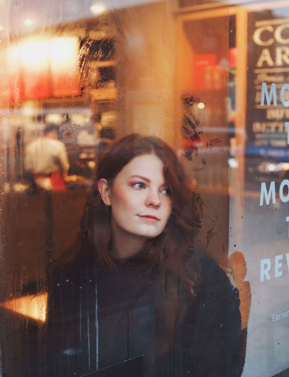 Moody portrait through window glass with cafe signage reflections in warm tones.