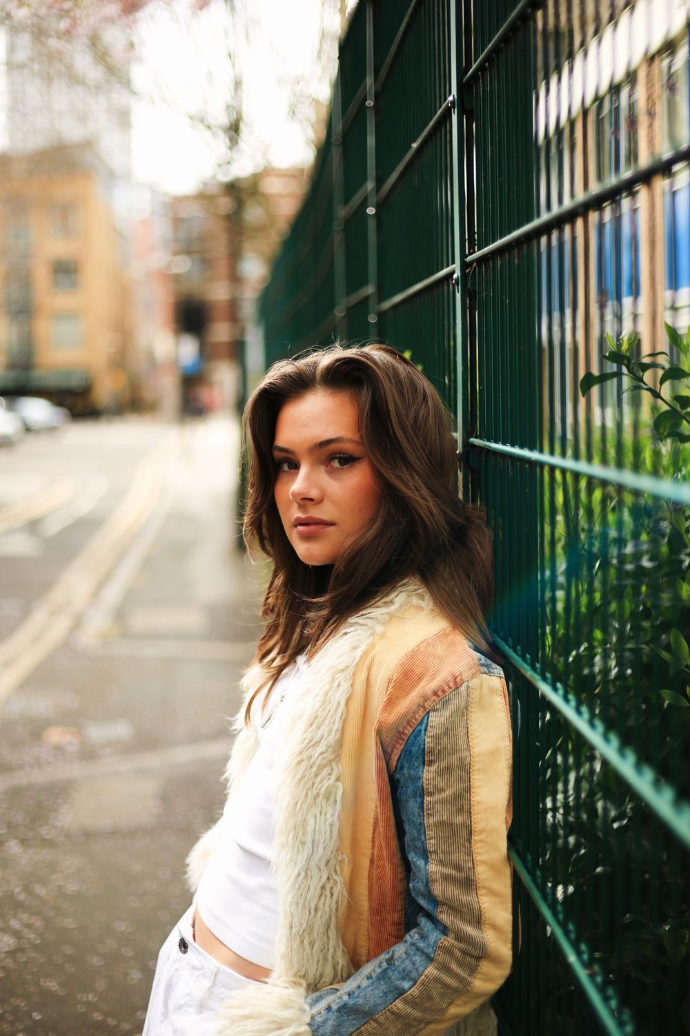 Casual fashion portrait of someone in a fur vest and jeans leaning against a green metal fence on a city street.
