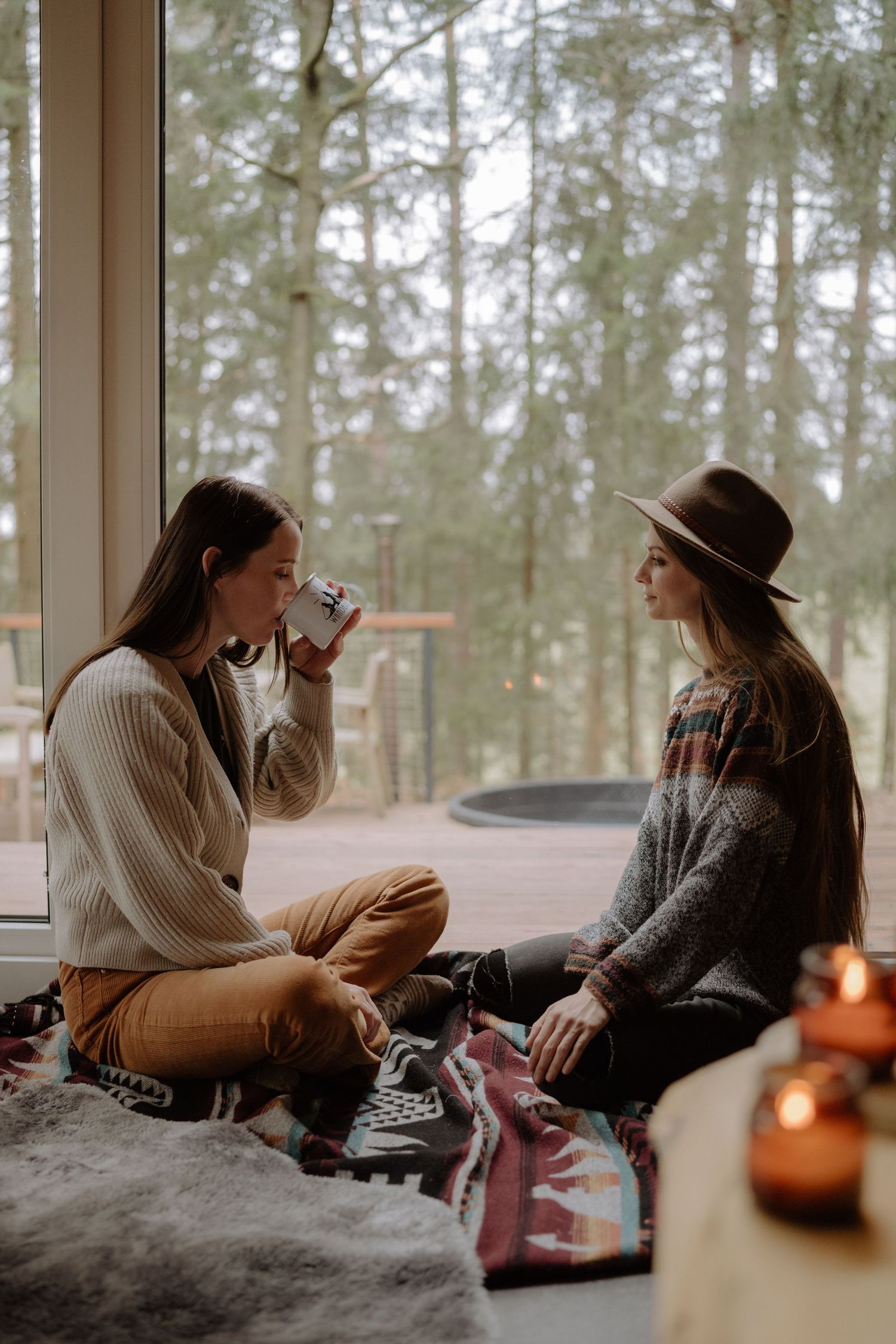 Two people share a cozy moment by large windows overlooking a forest.