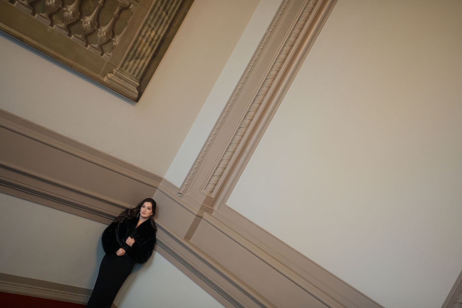 A person in dark clothing sits against a white wall with ornate molding details in a minimalist interior setting.