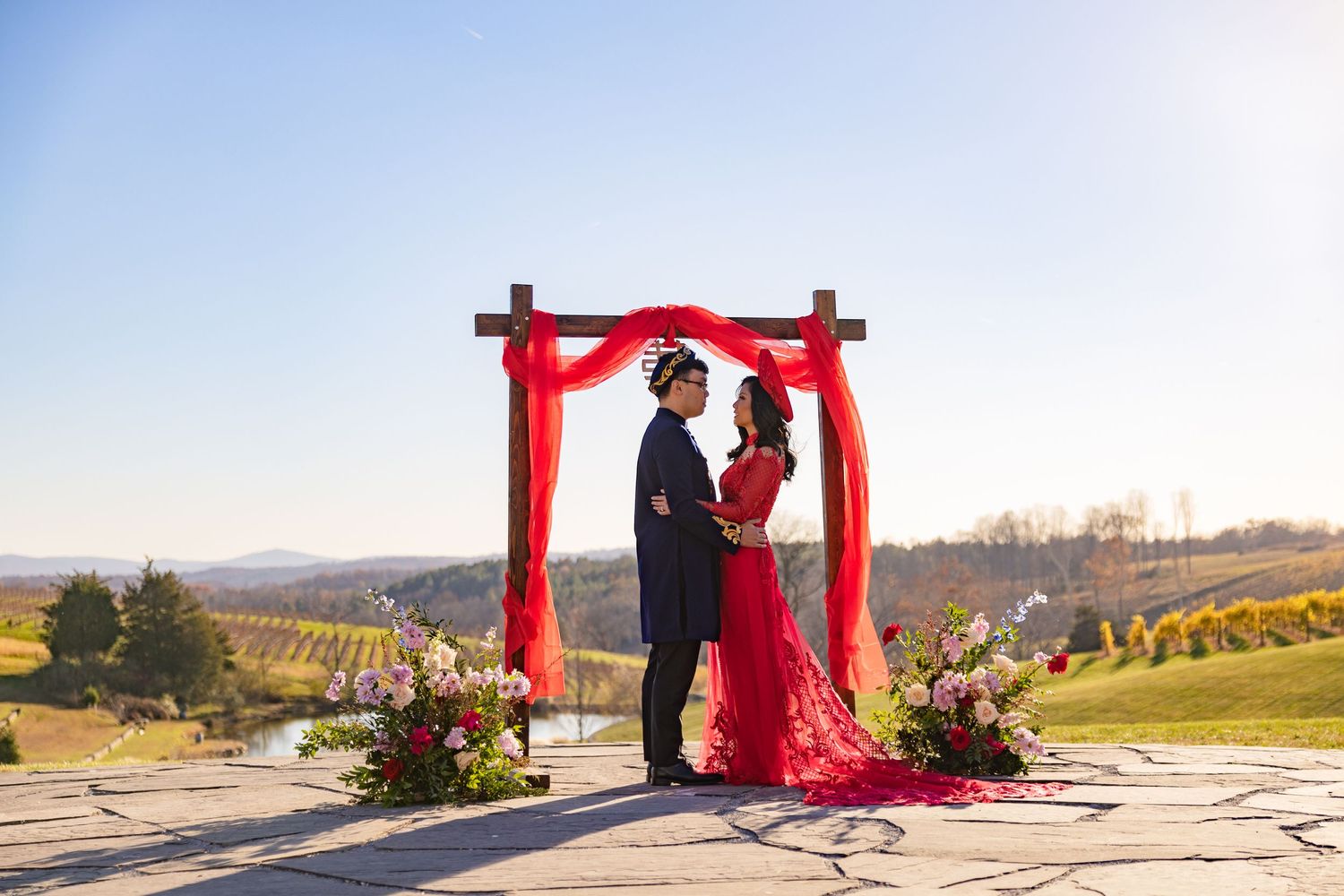 Asian wedding ceremony setup with red arch and floral arrangements against vineyard landscape.