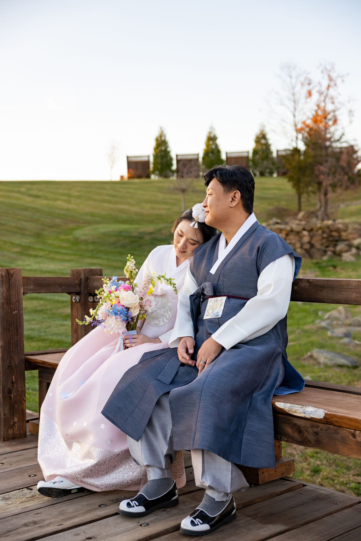 A couple in traditional Korean hanbok sits together on a wooden bench in a garden setting.