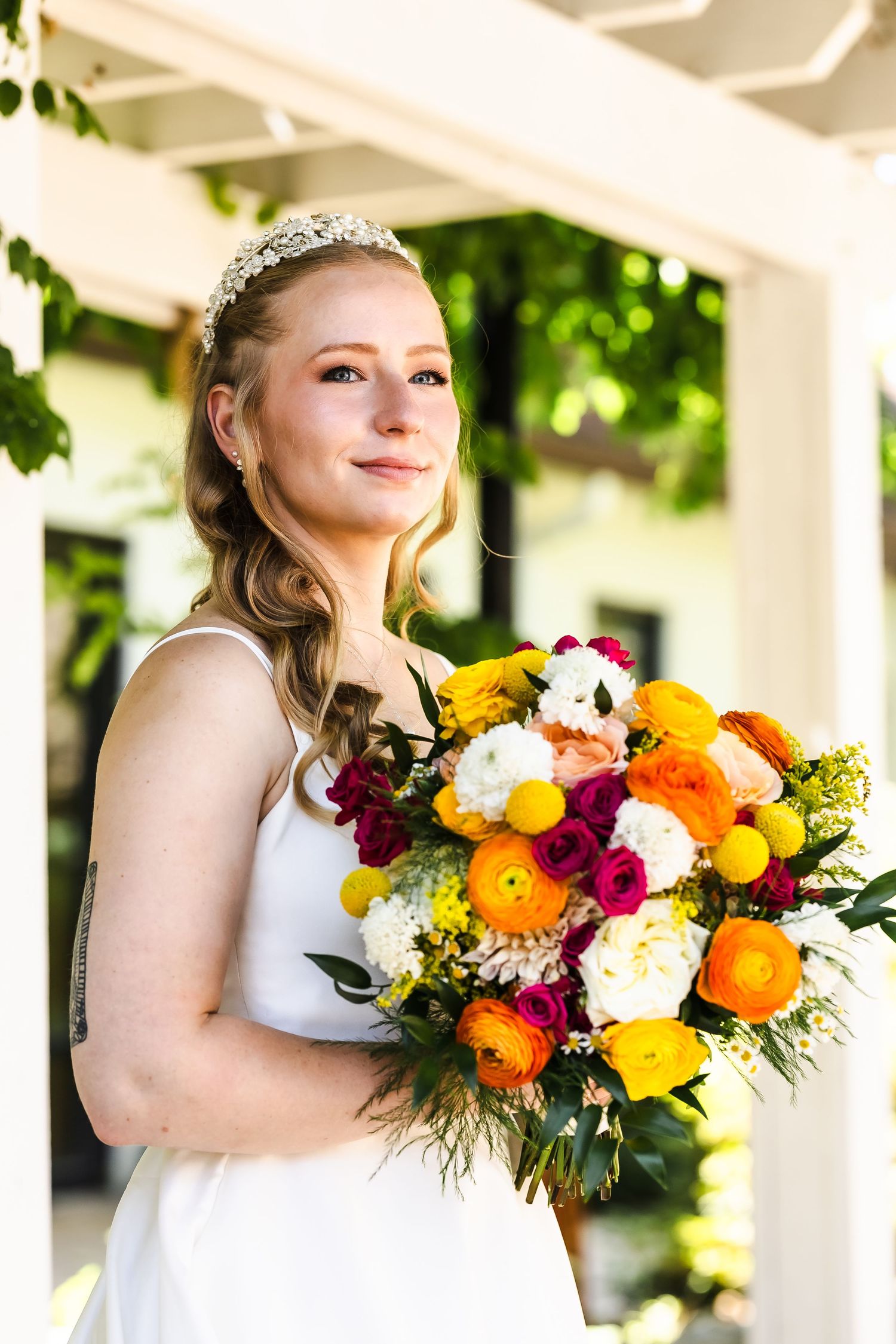 A bride holds a vibrant bouquet with orange, yellow and burgundy flowers against a white porch backdrop.