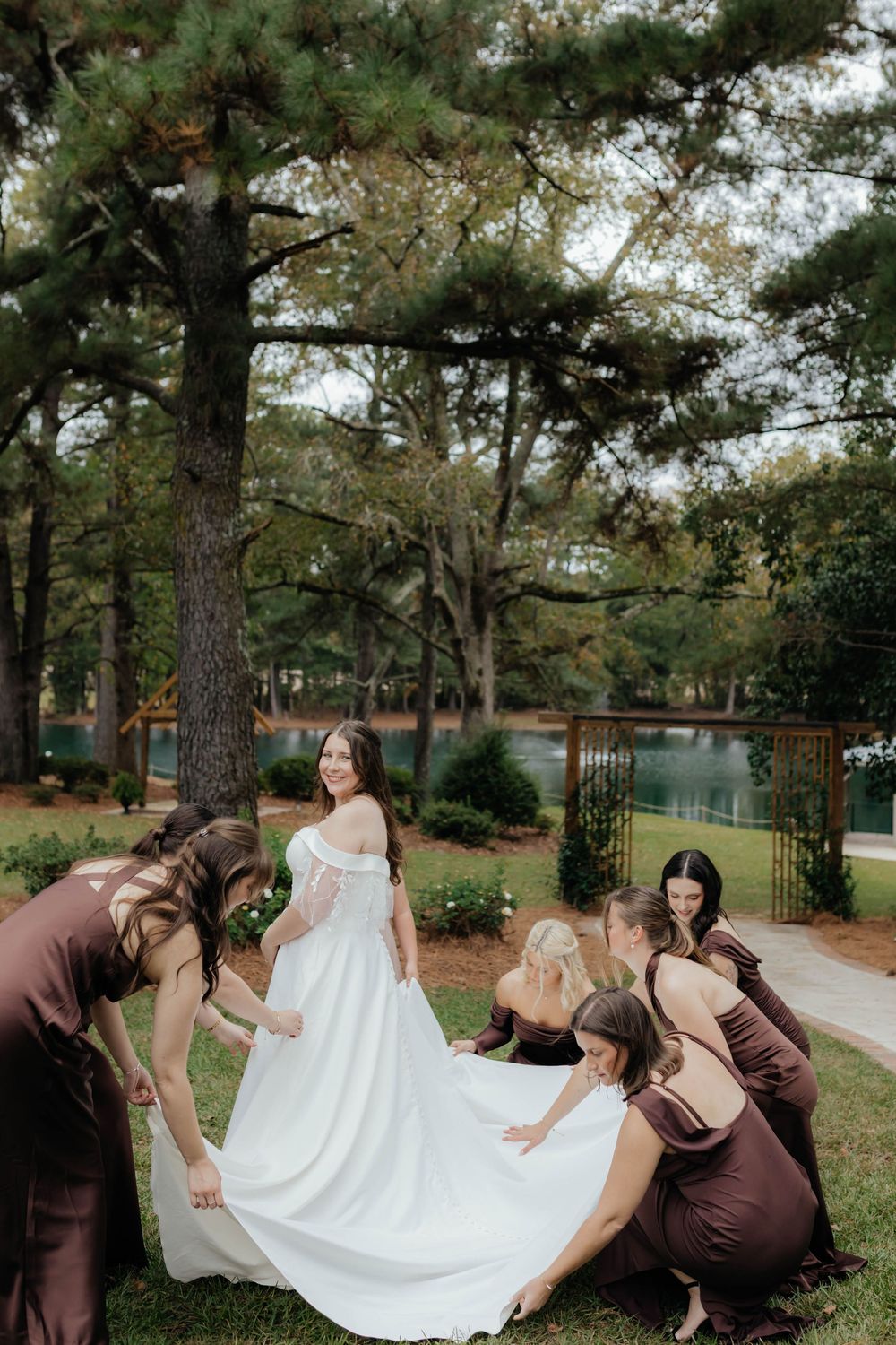 Bridesmaids in brown dresses help arrange the bride's wedding gown outdoors.