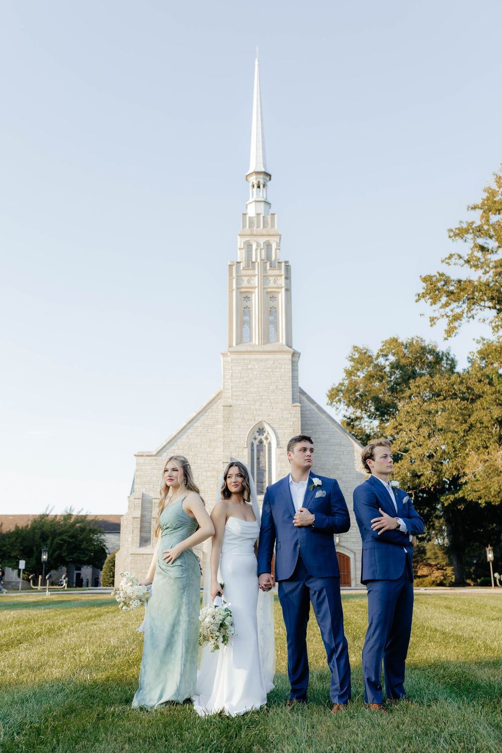 Wedding party poses on grassy hill with white stone church and steeple in background.