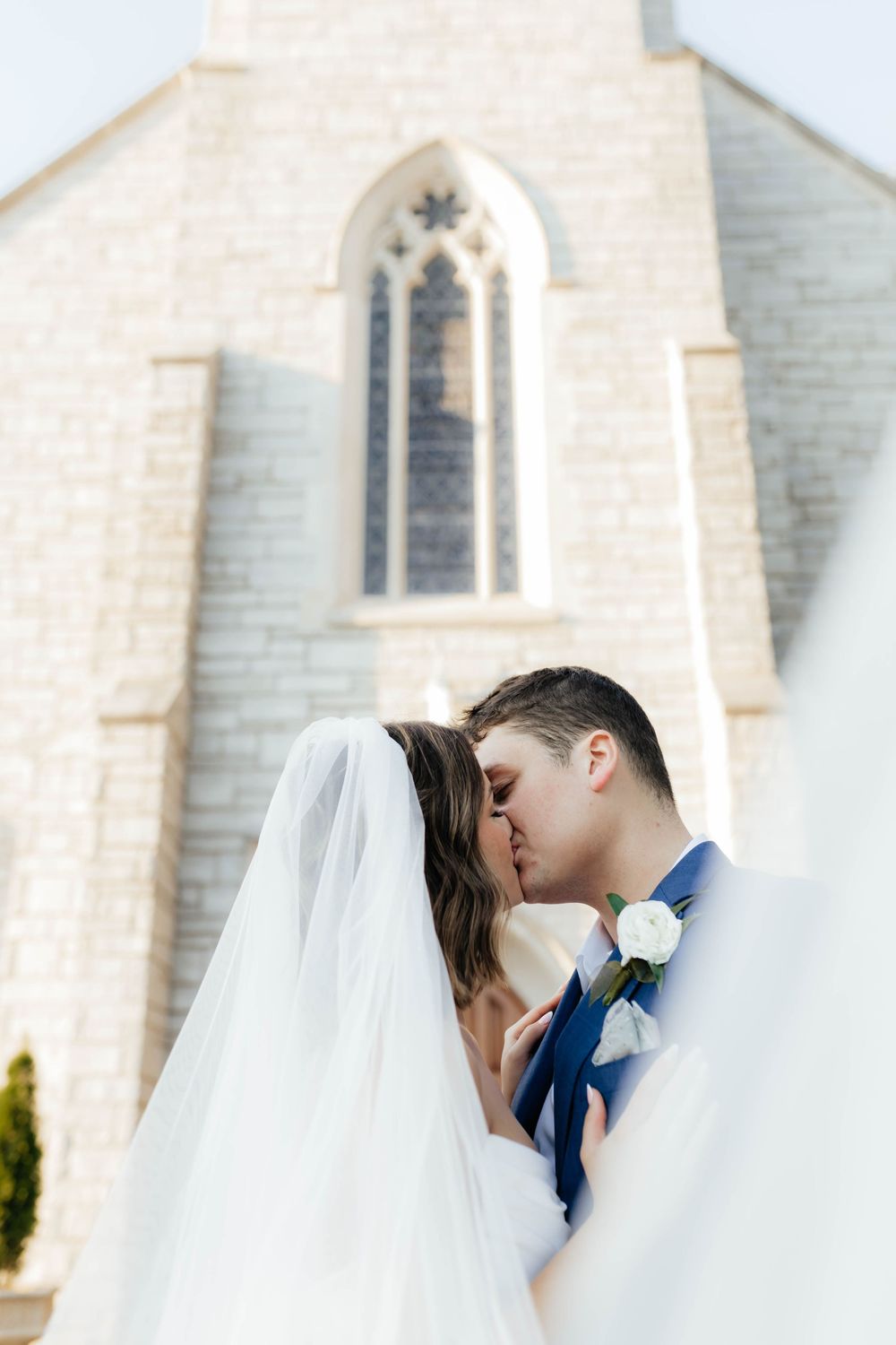 Romantic wedding kiss in front of a white stone church with gothic windows.