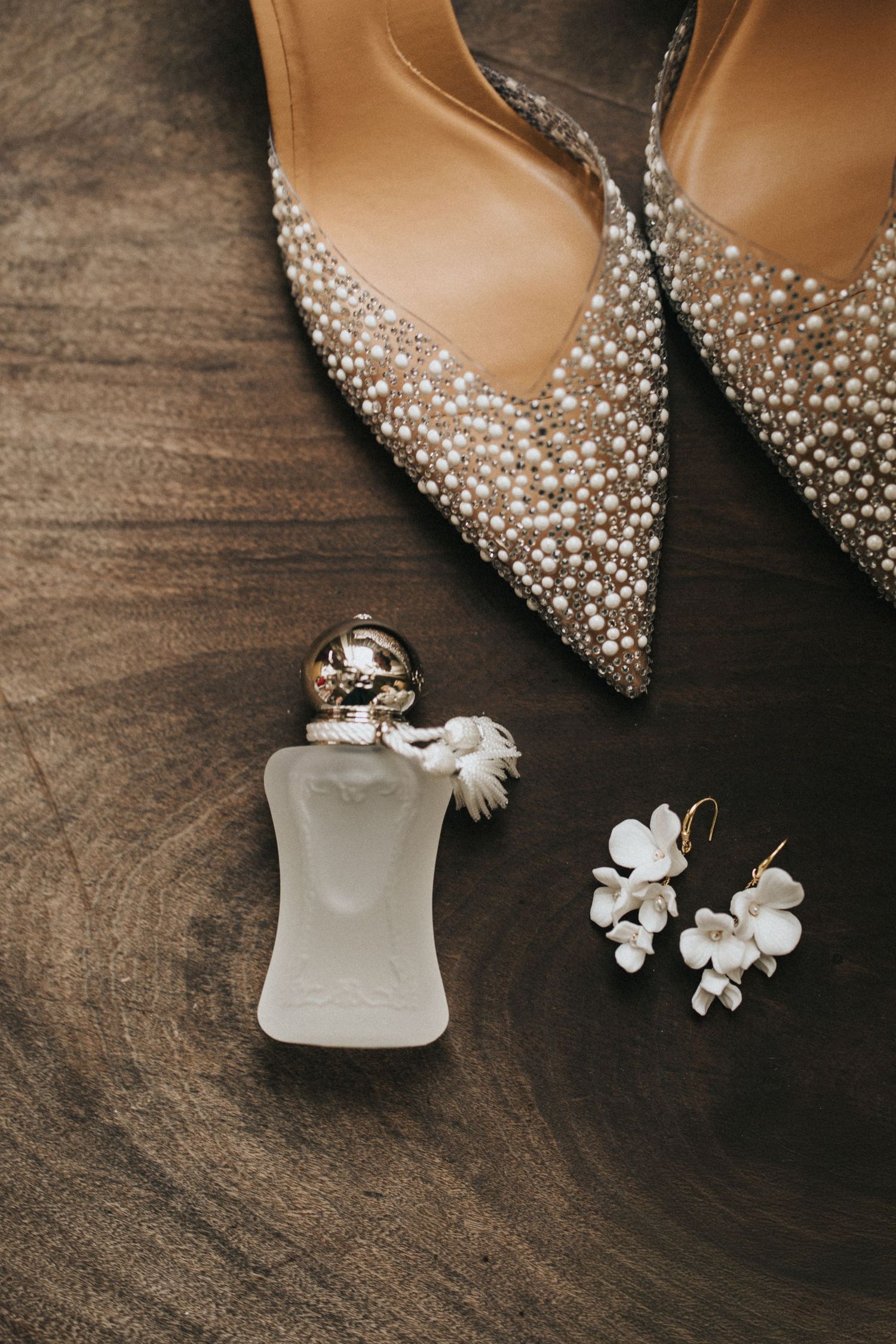 Elegant bridal accessories including sparkly pointed heels, a frosted perfume bottle, and delicate white floral earrings on wood.