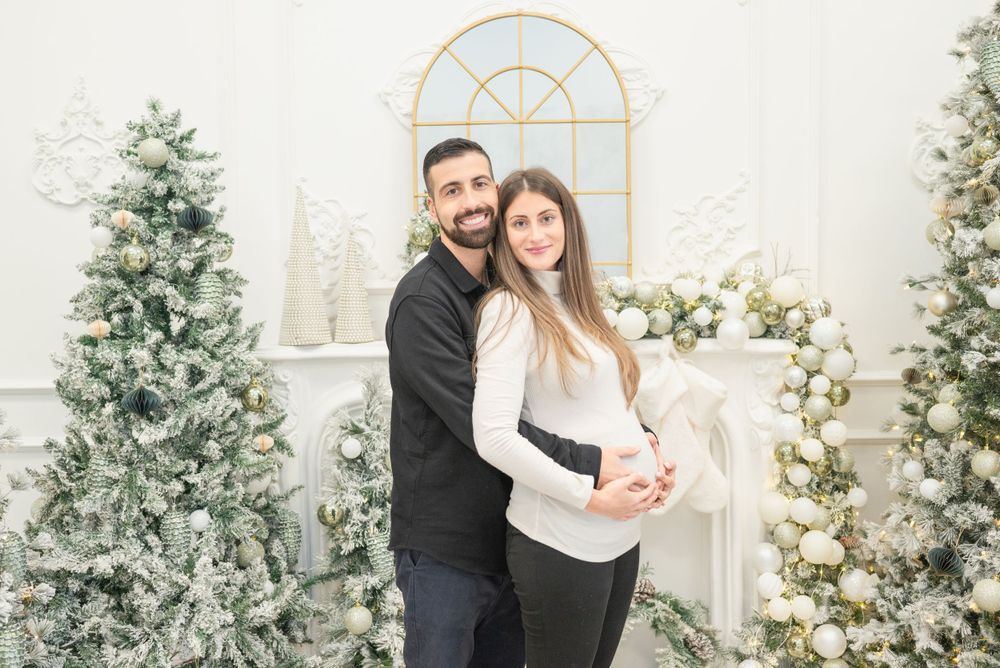 A couple poses together in an elegant winter setting with frosted Christmas trees and arched window.