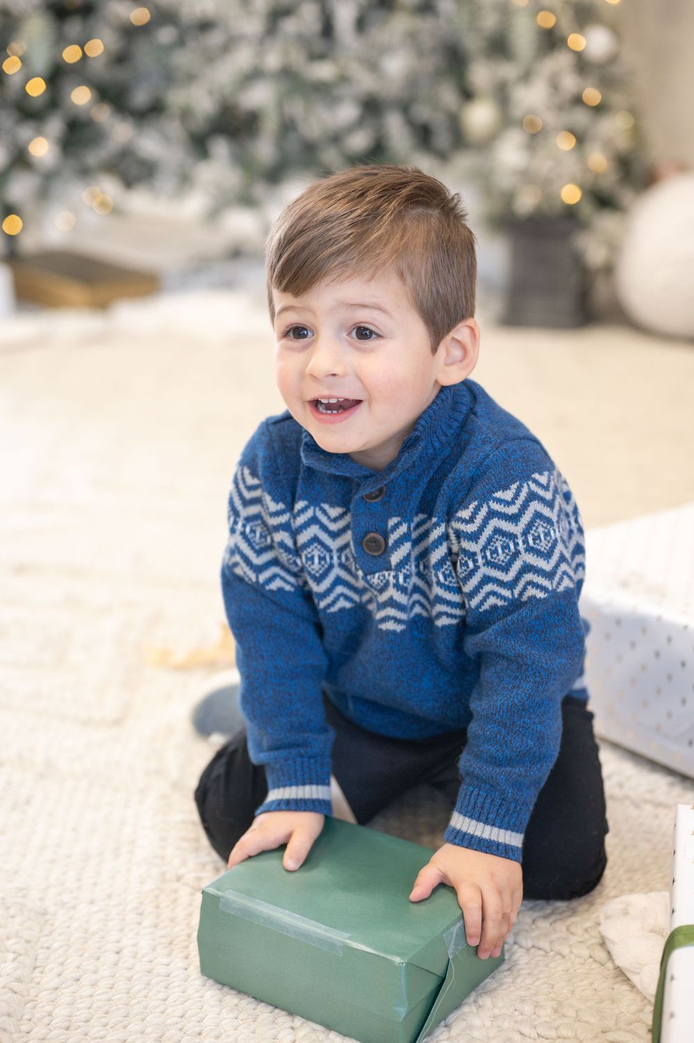 A child in a blue patterned sweater sits excited with a green wrapped present near a Christmas tree.