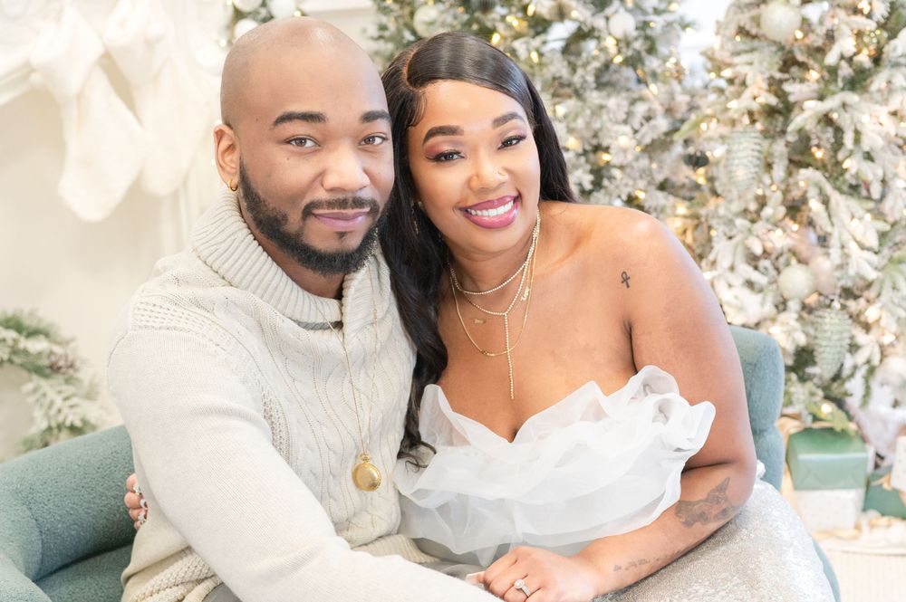 A couple poses together on a green couch with a decorated Christmas tree in the background.