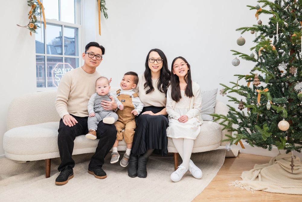 Family of five sitting together on white couch next to decorated Christmas tree.